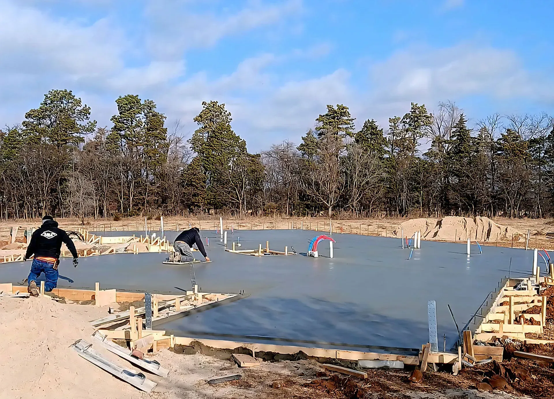 Workers pour concrete for a house foundation at a wooded construction site.