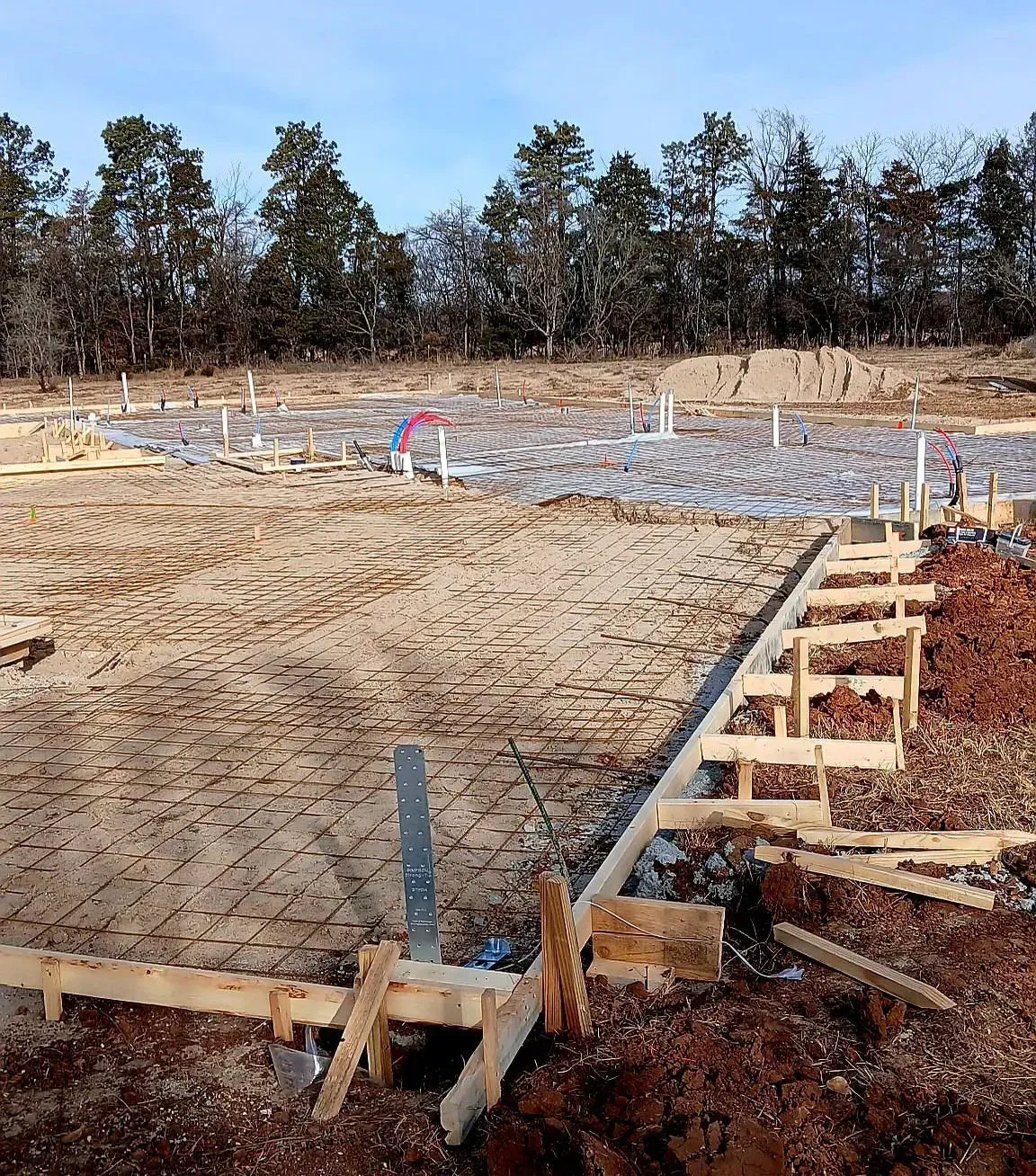 Foundation slab under construction with rebar, wooden forms, and workers on a dirt lot