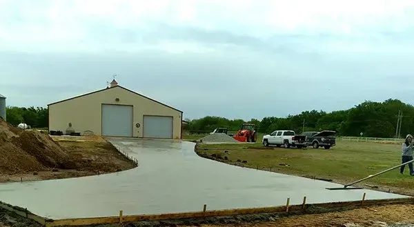 Concrete driveway leading to a beige warehouse with a garage door and parked trucks on a cloudy day