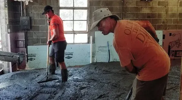 Two workers in orange shirts shovel wet concrete inside a dim construction room.