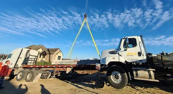 White flatbed truck lifting a trailer with yellow straps under a blue sky on a work site