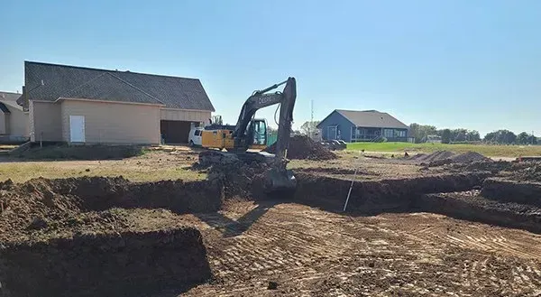 Excavator digging earth at a residential construction site beside new houses under a clear sky.
