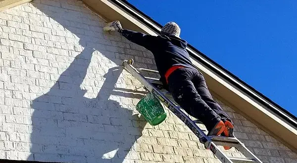 Person on a ladder painting a house exterior under a blue sky