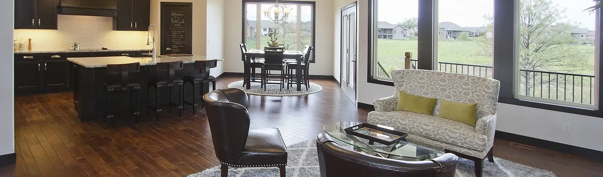 Bright open-plan living room and kitchen with dark wood floors, black stools, and a sofa by large windows.