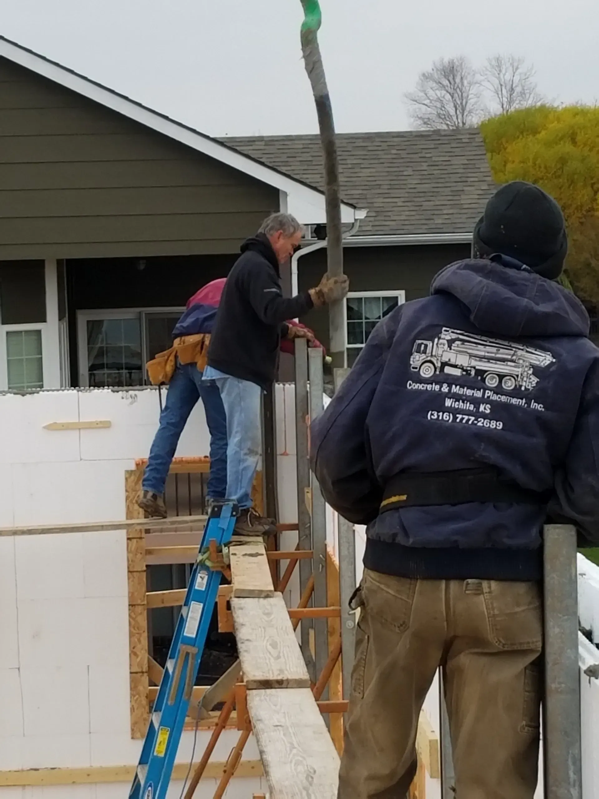 Two workers using ladders to install a tall pole beside a house
