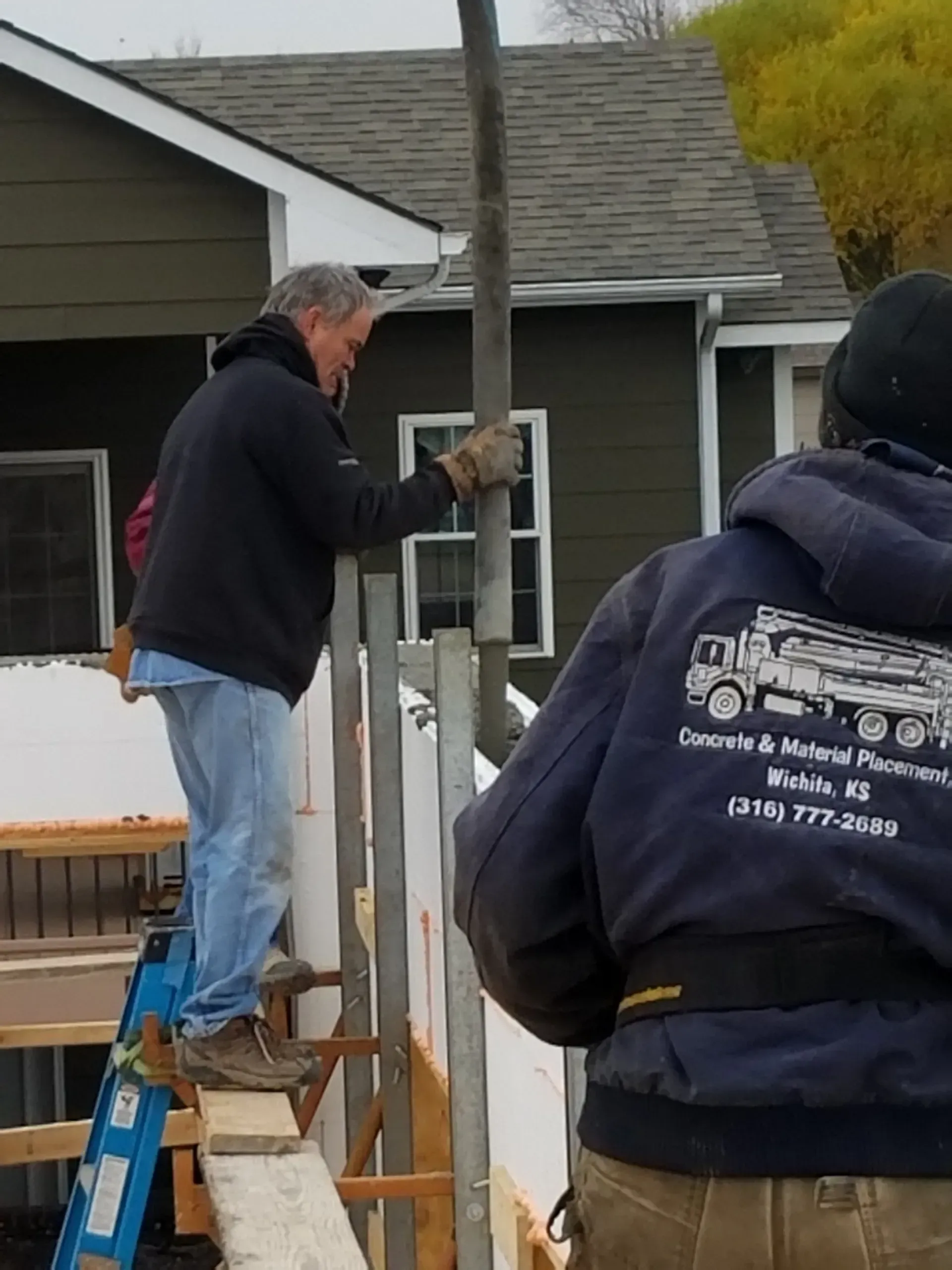 Worker on a blue ladder installing metal hardware on a porch post while another person stands nearby.
