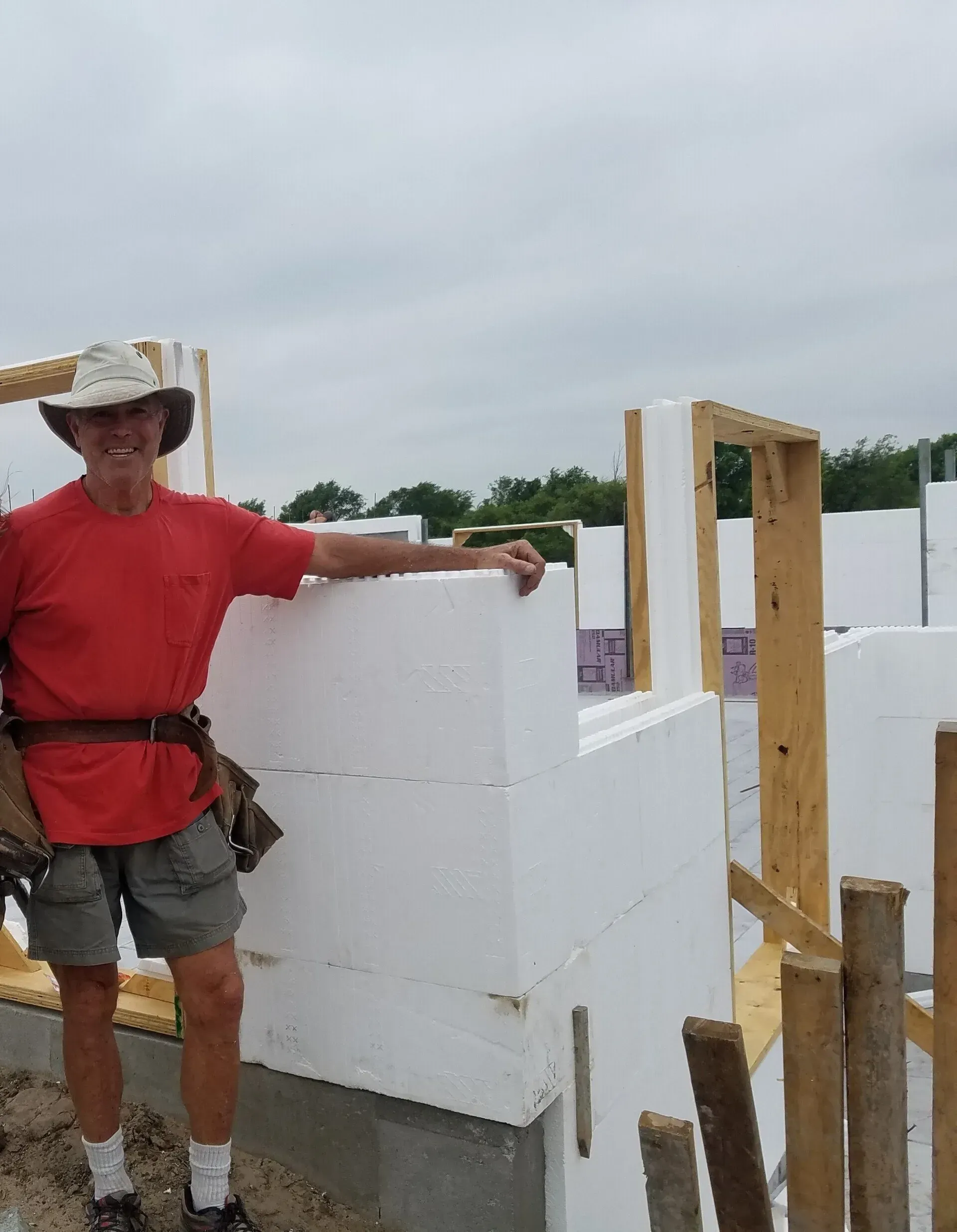 Man in red shirt and cowboy hat standing beside a partially built white wall at a construction site