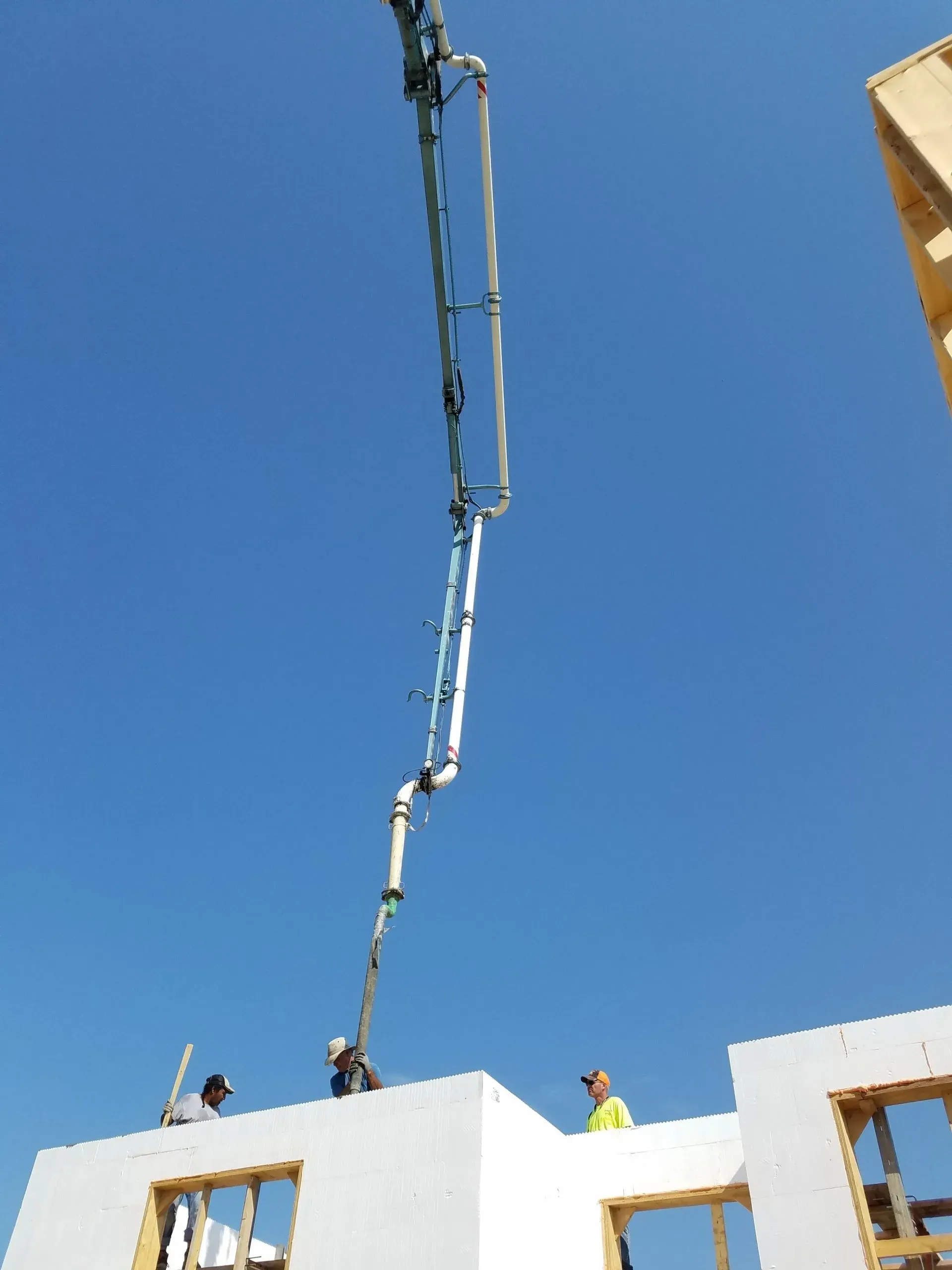 Workers using a long concrete pump to pour a roof on a white building under a clear blue sky