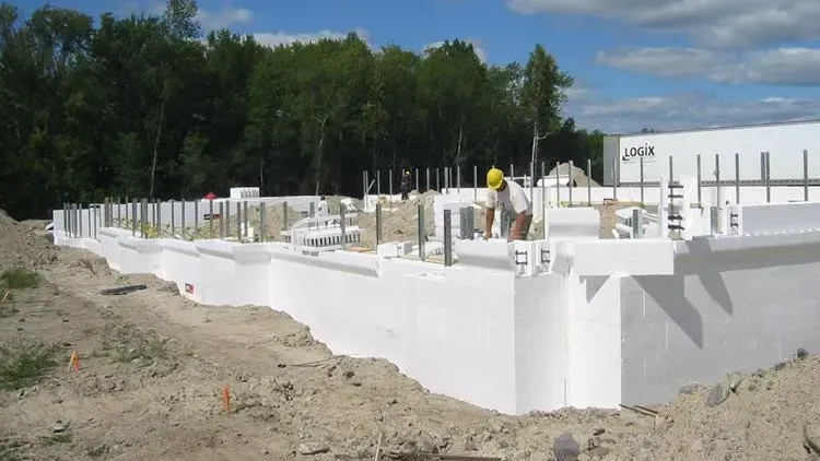 White concrete foundation walls under construction on a dirt lot beside a wooded area