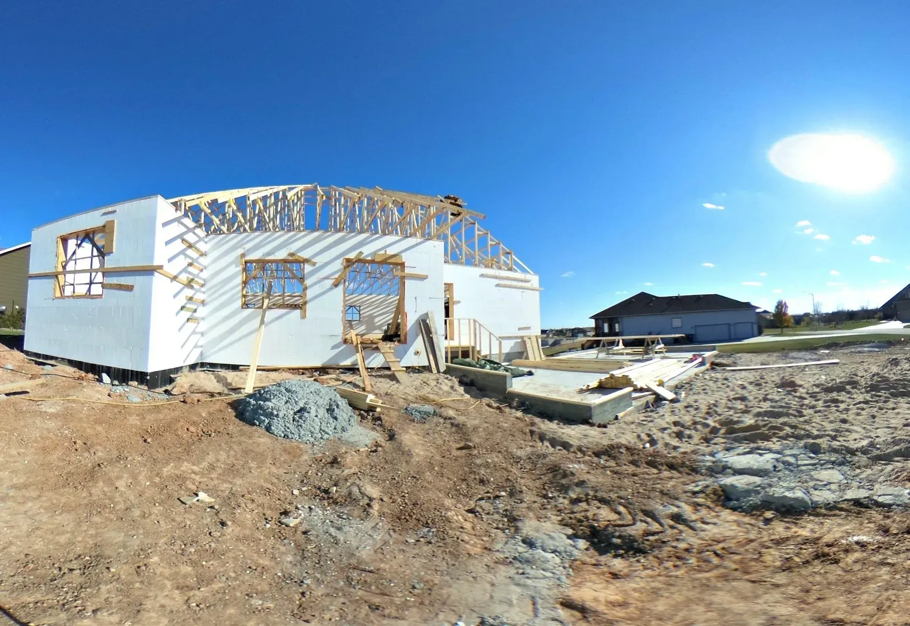 House under construction with white siding, exposed framing, and a bright blue sky over a muddy lot