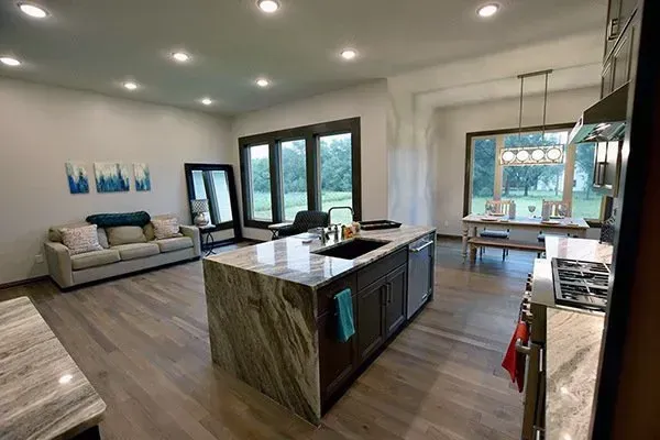 Modern open-plan kitchen and living room with island, gray sofa, and large windows overlooking greenery