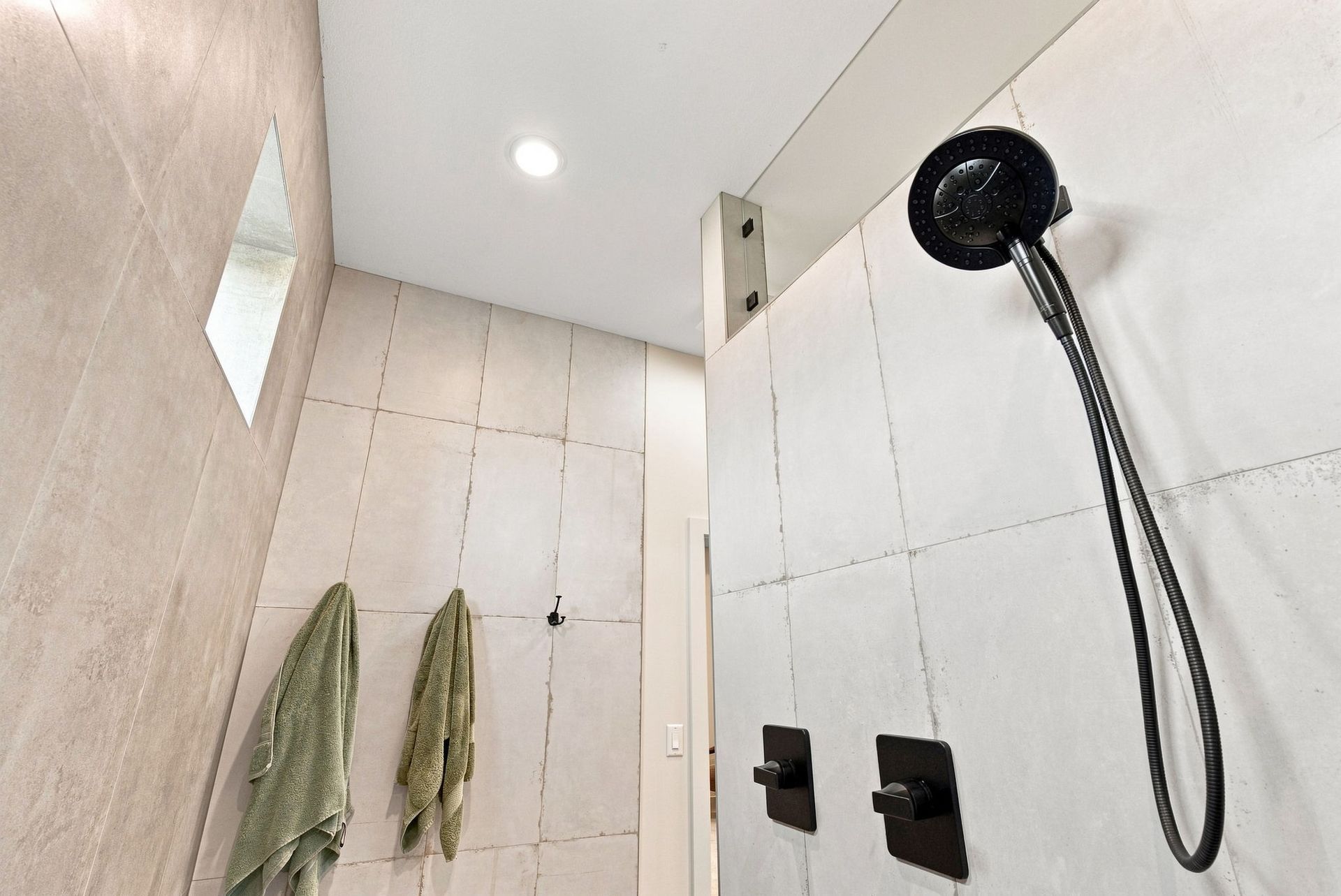 Modern shower with white tiled walls, black showerhead, and hanging towels under a ceiling light