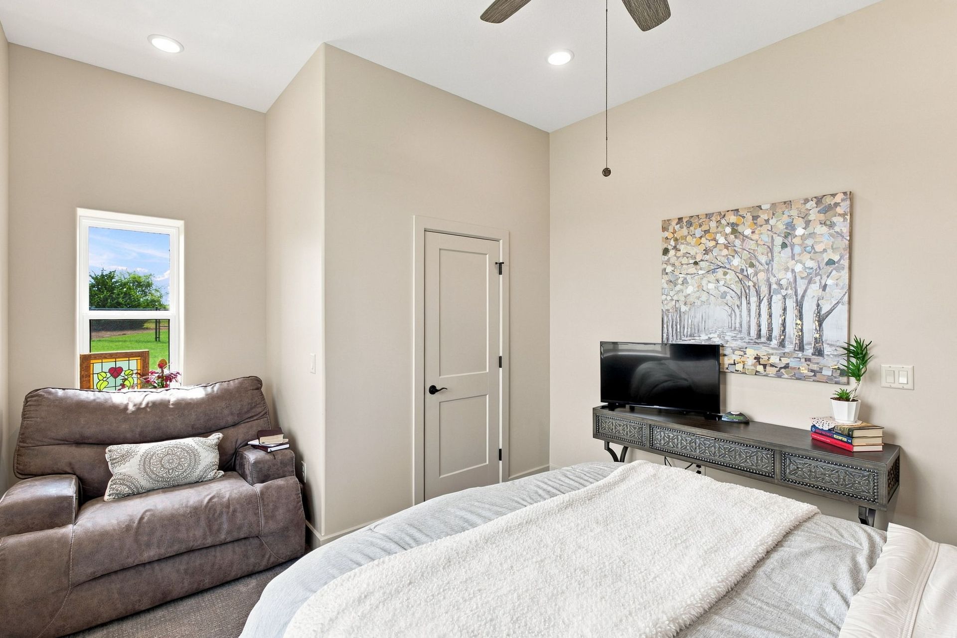 Bright bedroom with a bed, armchair, TV stand, and artwork on beige walls