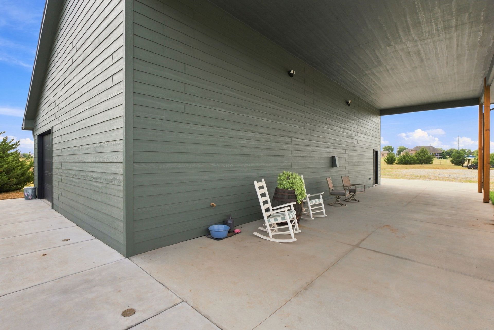 Covered patio beside a gray house with white chairs, a small table, and potted plants on concrete floor