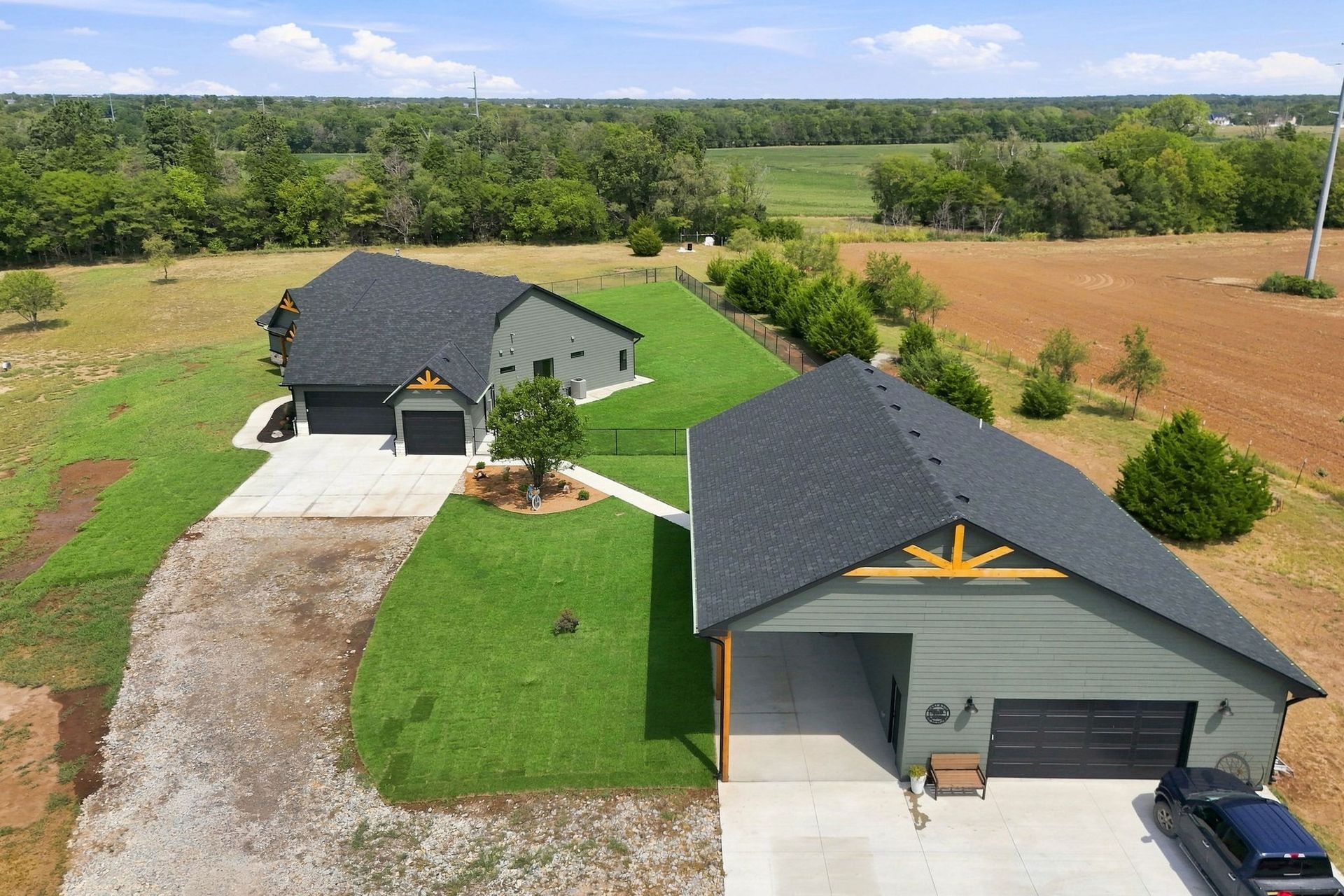 Aerial view of two dark-roofed houses with long driveways, green lawns, and surrounding trees in a rural area