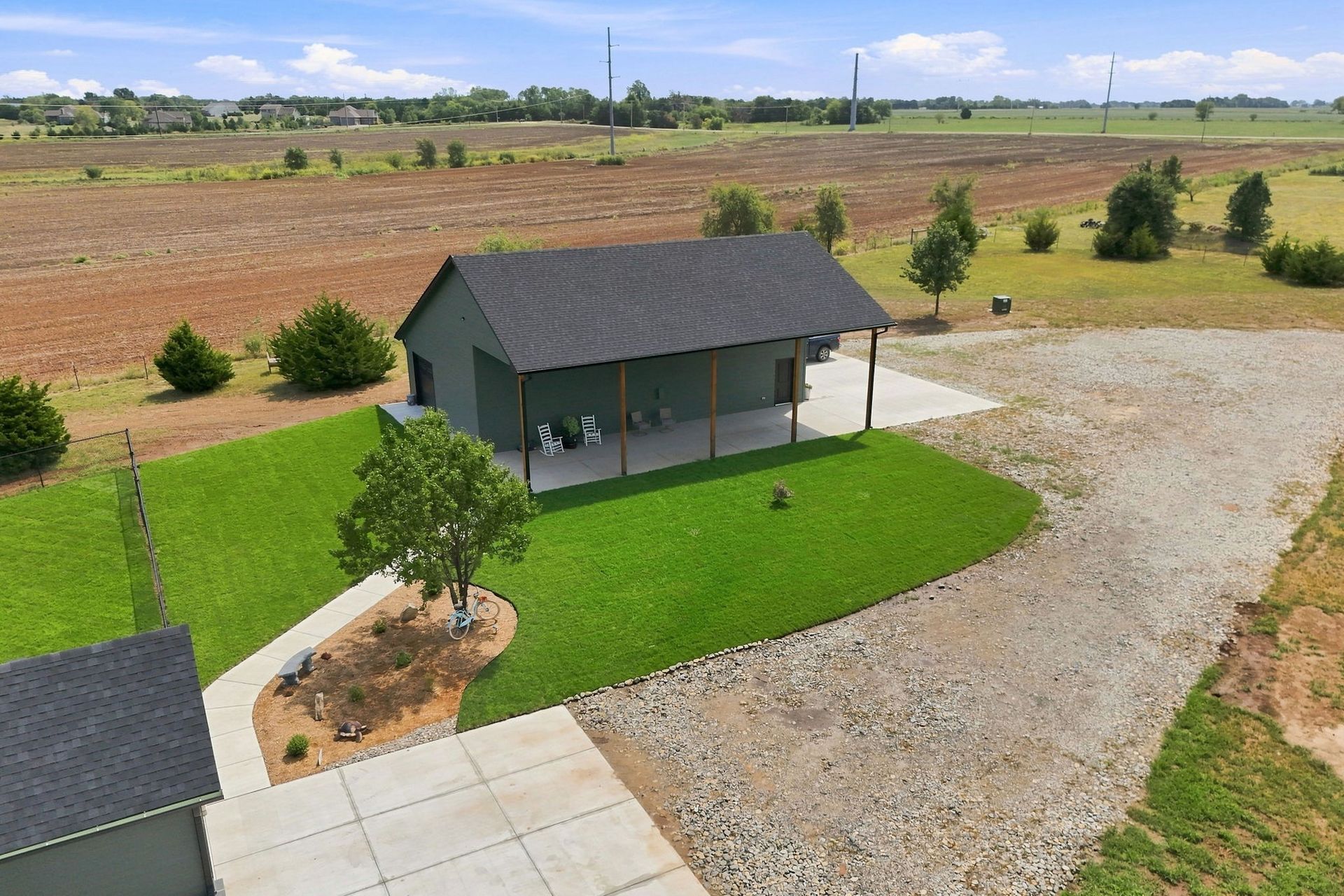 Small gray shed on a grassy lot with a gravel driveway, viewed from above, with fields in the background