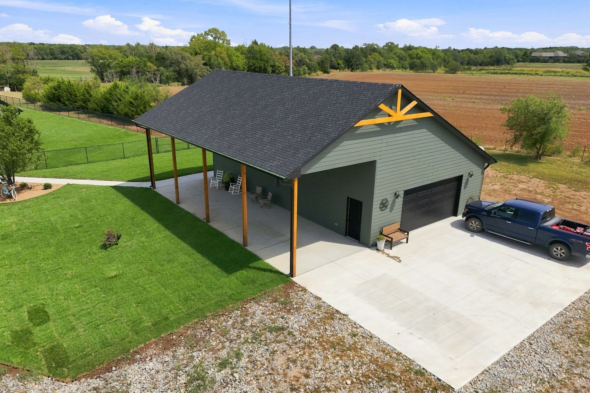 Small gray outbuilding with black roof, covered porch, and parked car beside a concrete driveway in a rural setting