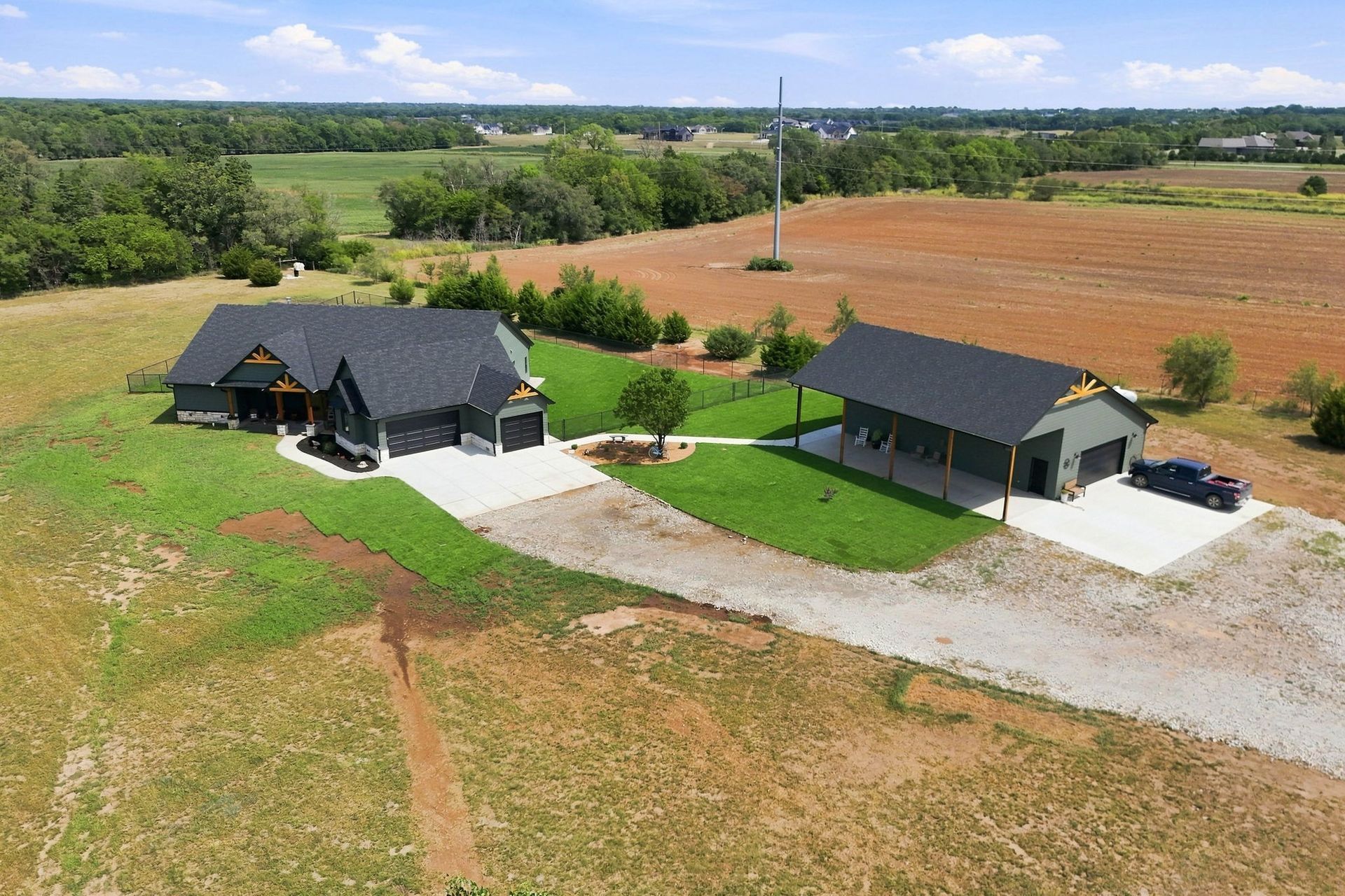 Two dark-roofed houses on a rural property with gravel driveways and open fields beyond.
