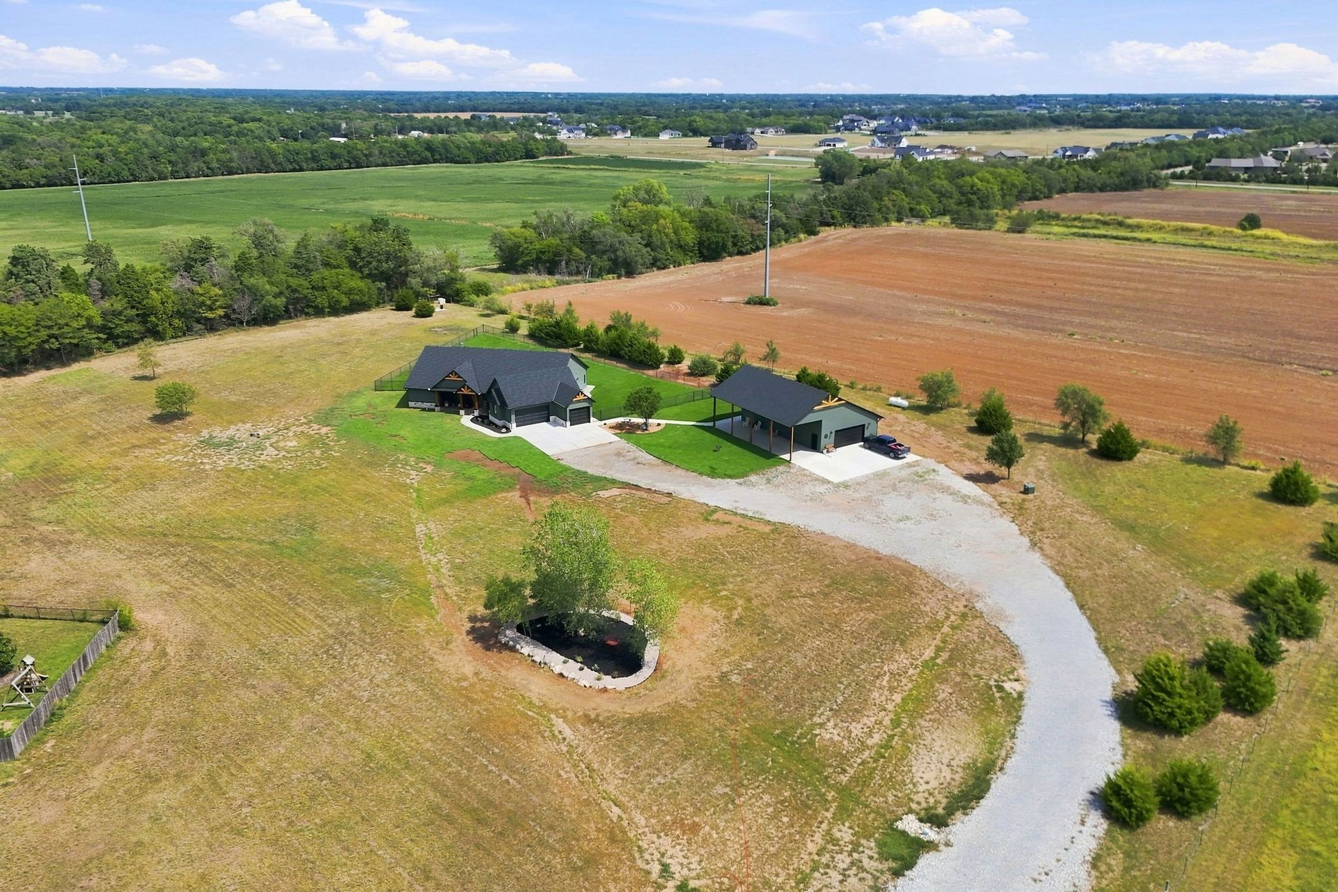 Aerial view of a rural house with a curved driveway, open fields, and trees under a clear sky