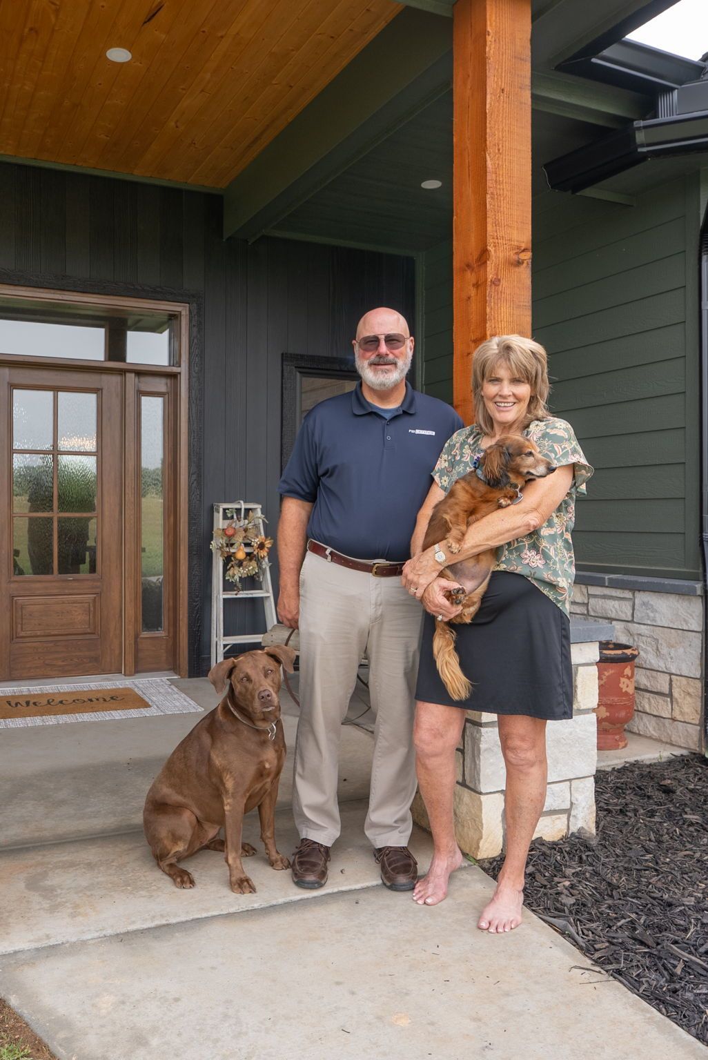 Couple standing on a front porch with a brown dog; woman holding a tabby cat.