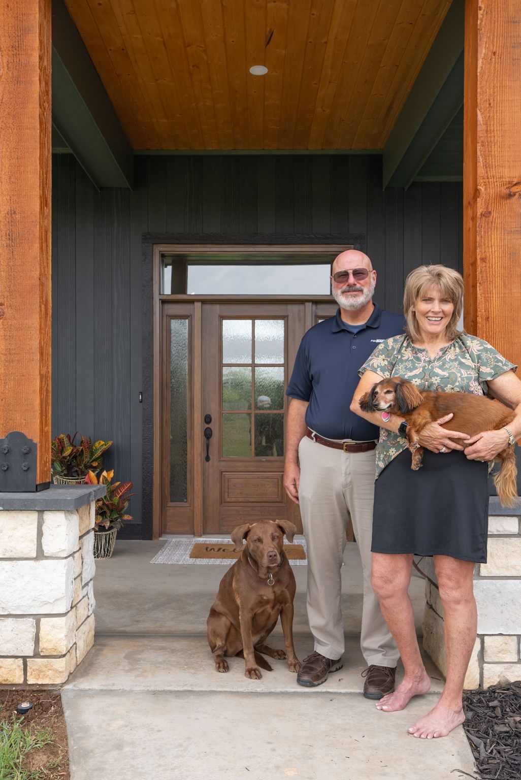 Couple standing on a porch with two brown dogs, one held in arms, in front of a front door