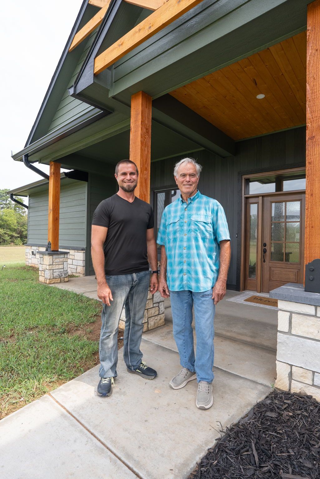 Two men standing on a front porch of a modern house, one in black and one in blue plaid.