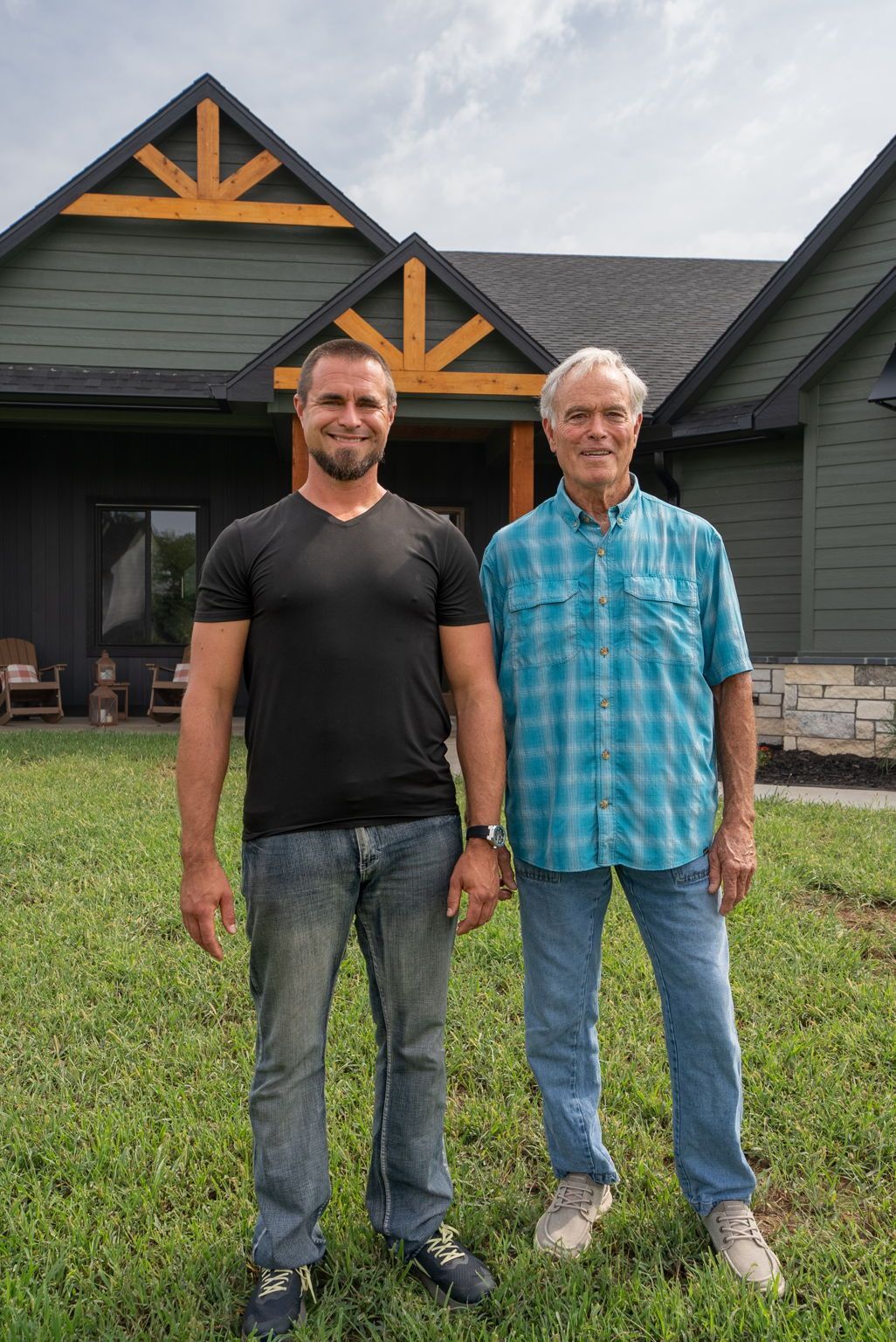 Two men standing on grass in front of a dark house, one in a black T-shirt and one in a blue plaid shirt