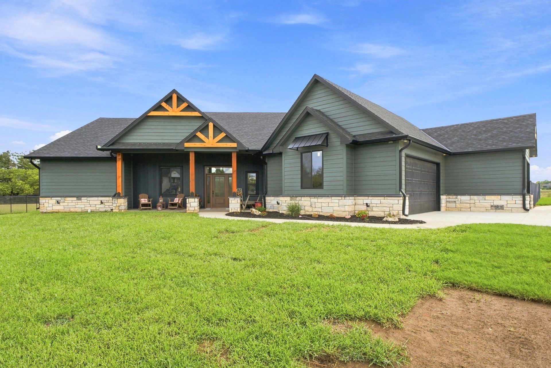 Gray house with stone base, dark roof, and orange trim, set on a grassy lot under a blue sky