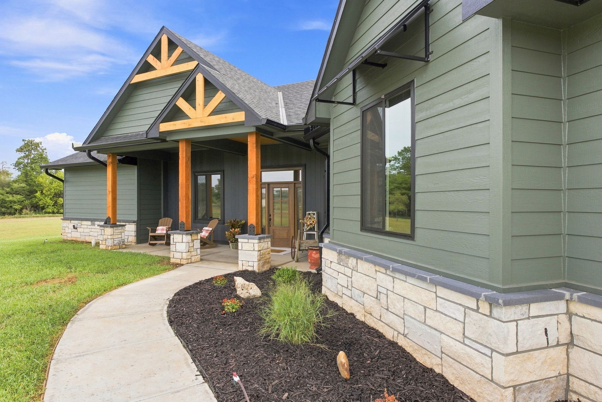 Covered porch of a green house with stone base, wood trim, and a curved walkway in a grassy yard