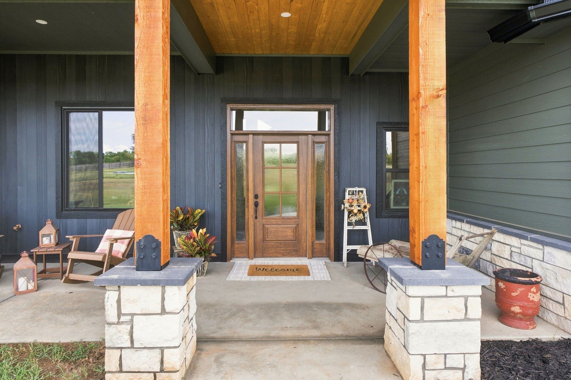Front porch with stone pillars, wooden ceiling, and glass front door, flanked by chairs and potted plants