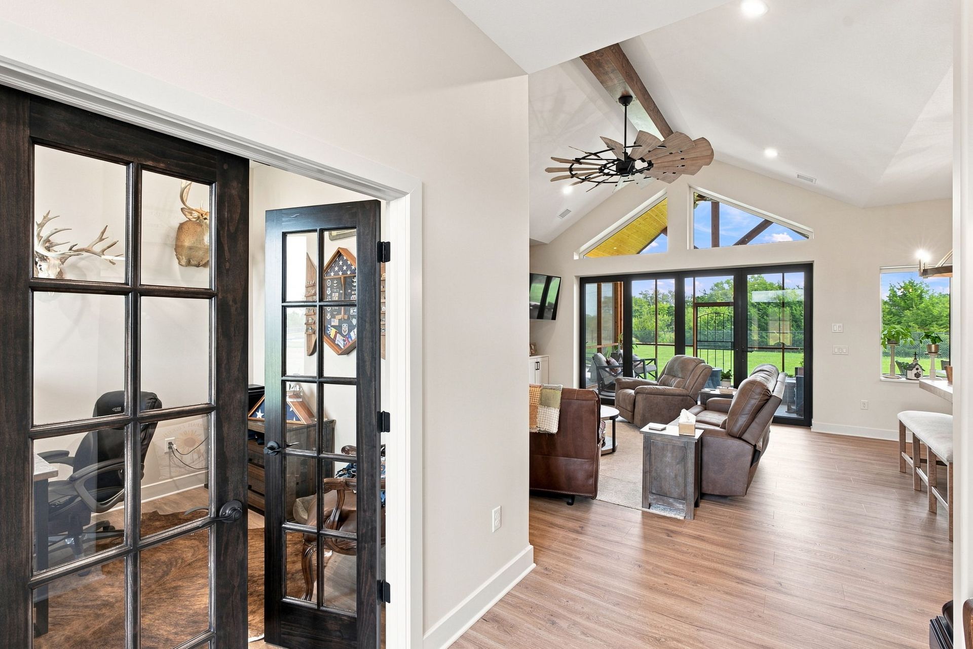 Bright open living room with hardwood floors, black-framed glass doors, ceiling fan, and gray seating by large windows