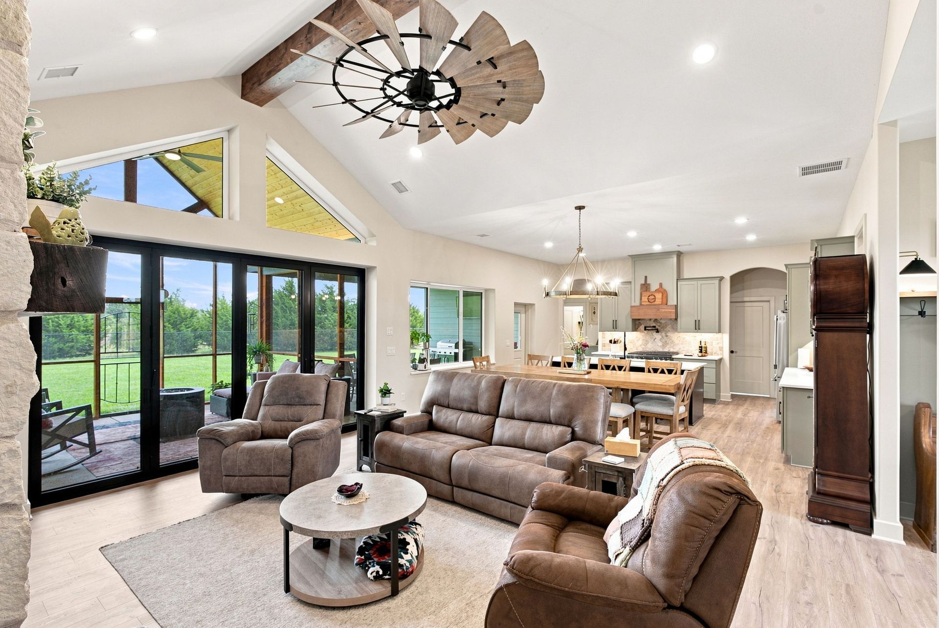 Bright open-concept living room with brown sofas, dining area, and large glass doors overlooking a green yard