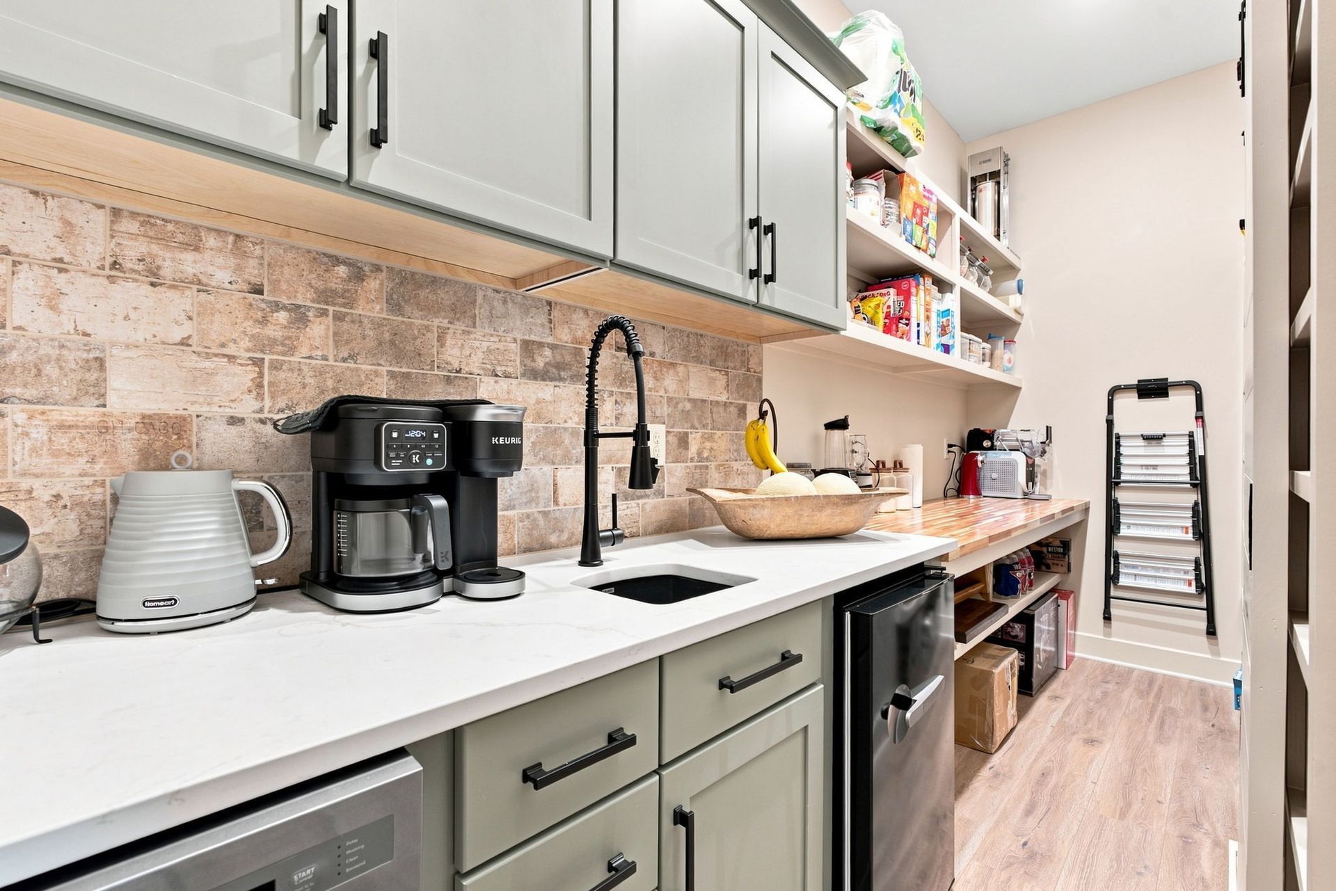 Compact kitchen or pantry with white cabinets, coffee maker, sink, and open shelving leading to a hallway.