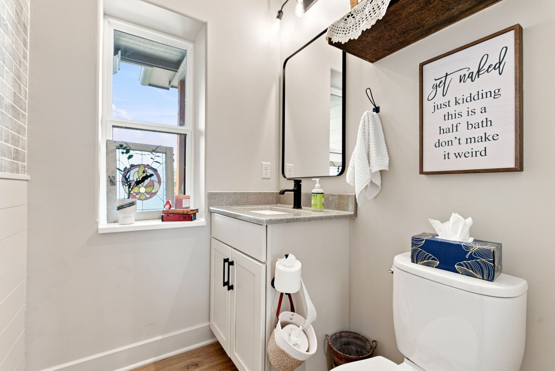 Small white bathroom with vanity, toilet, window, and framed wall sign.