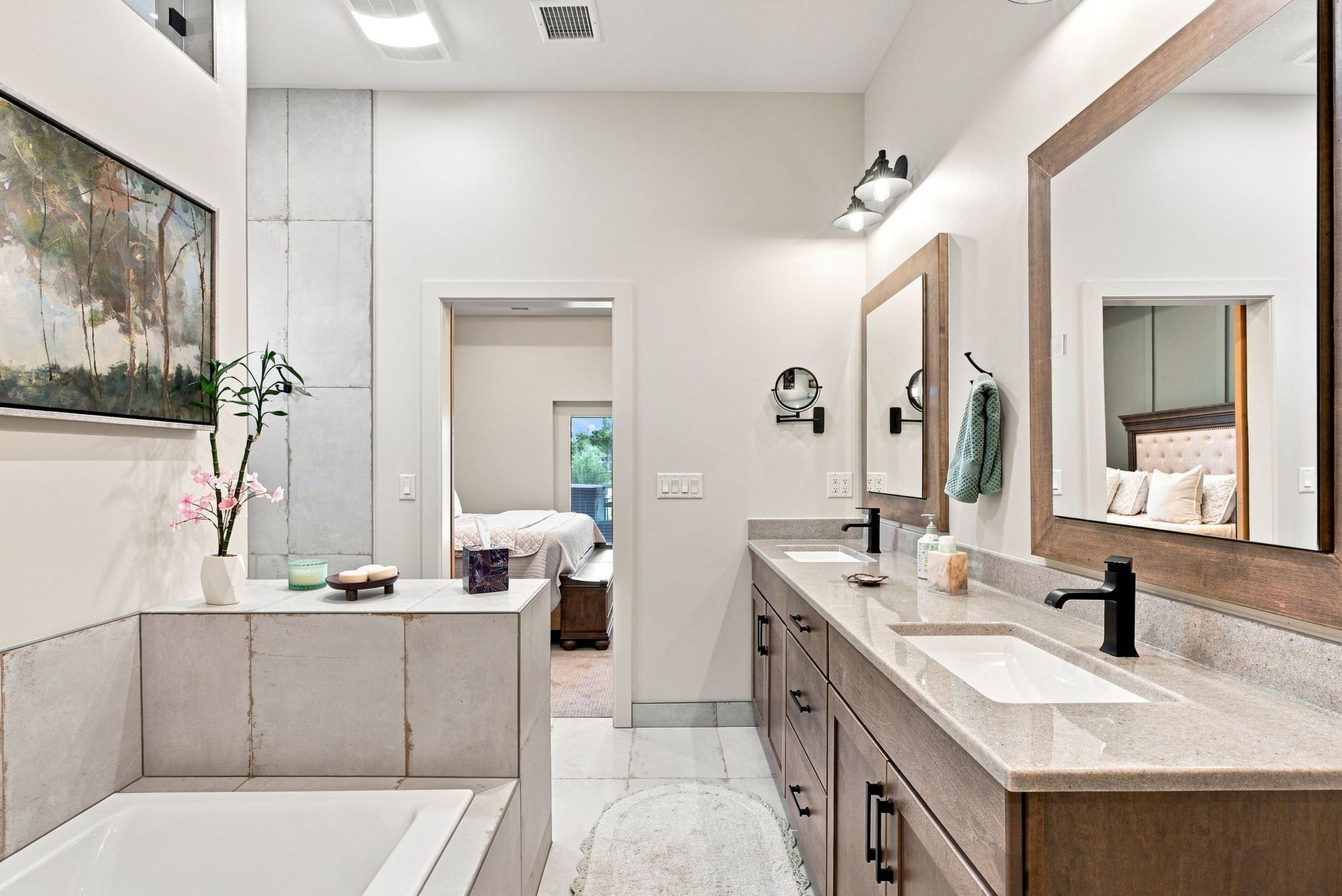Bright modern bathroom with double vanity, large mirror, soaking tub, and doorway to a bedroom.