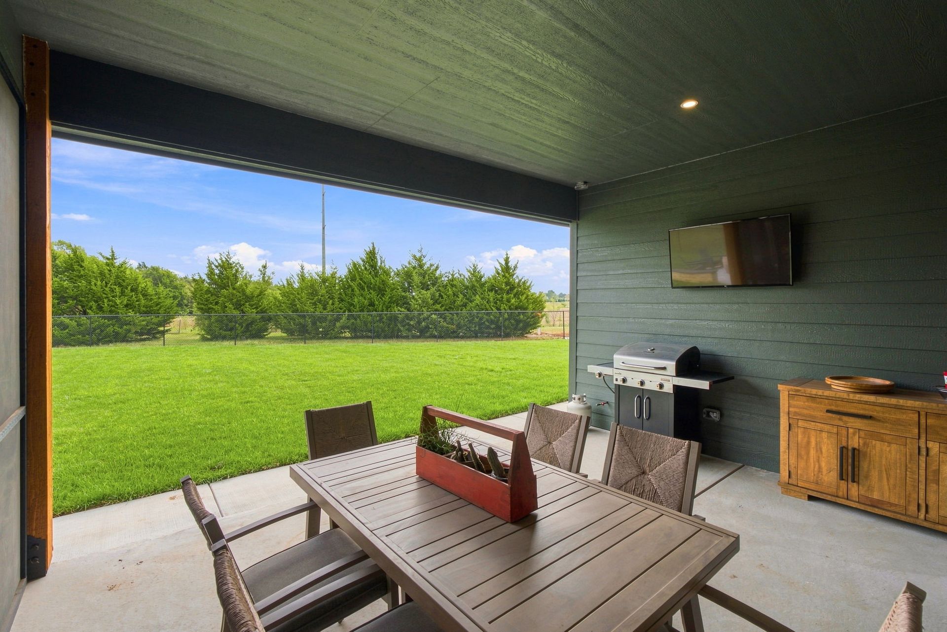 Covered patio with dining table, grill, and TV overlooking a green lawn