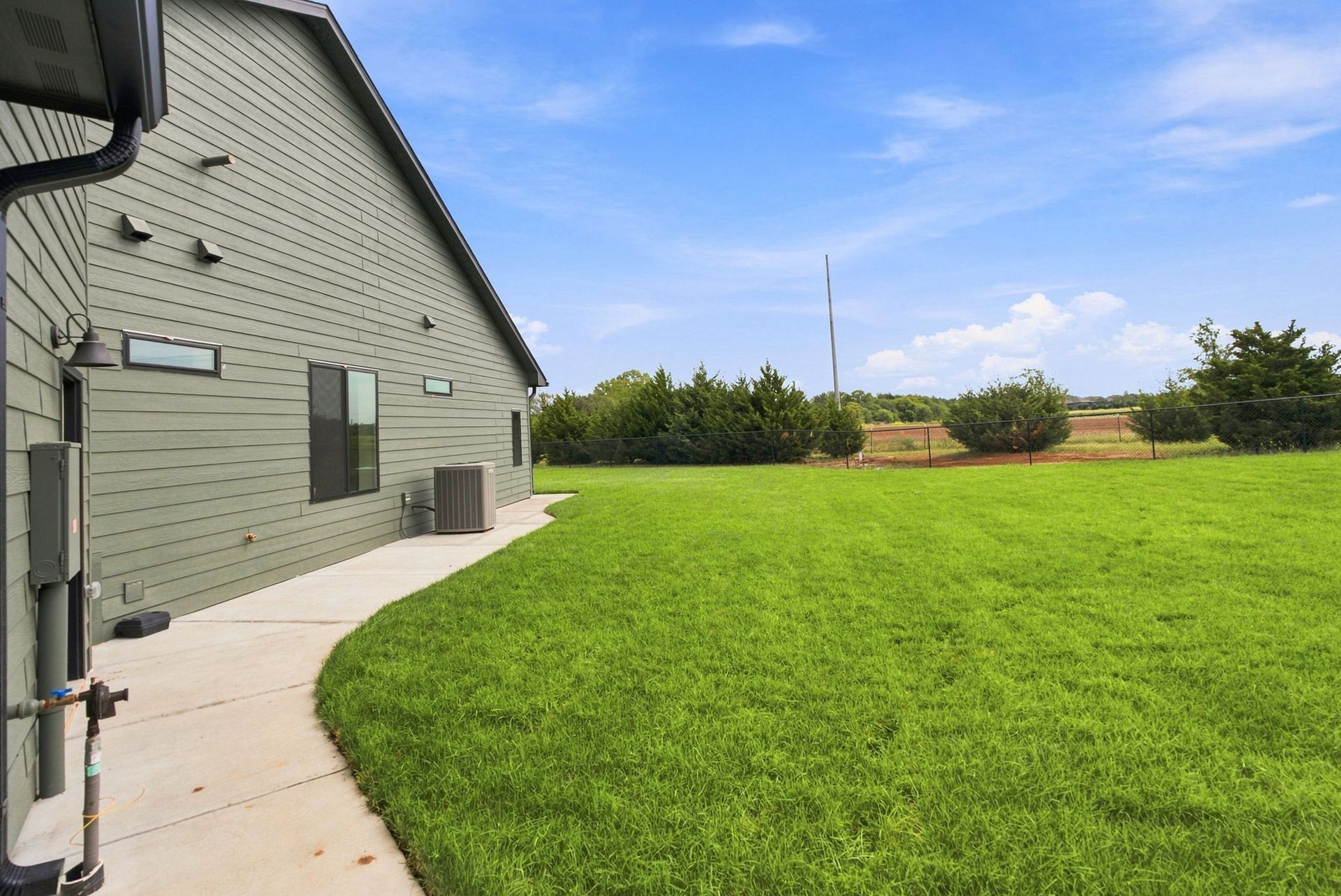 Backyard beside a gray brick house with a curved patio and a wide green lawn under a blue sky