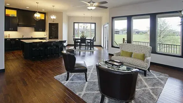Modern open-plan living room with dark wood floors, kitchen island, and large windows overlooking a green field