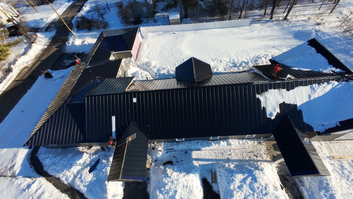 Aerial view of a snow-covered dark roof with chimneys and surrounding trees in winter.