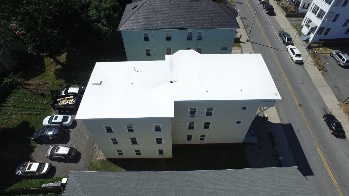 Aerial view of a white apartment building beside a road with parked cars and trees.