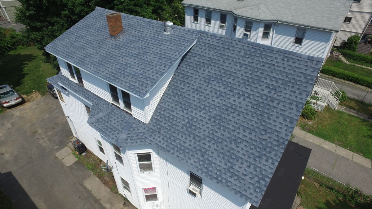 Aerial view of a white house with a blue shingled roof and dormer windows near a driveway and sidewalk