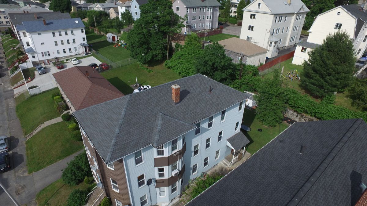 Aerial view of apartment buildings with gray roofs and surrounding trees in a residential neighborhood.