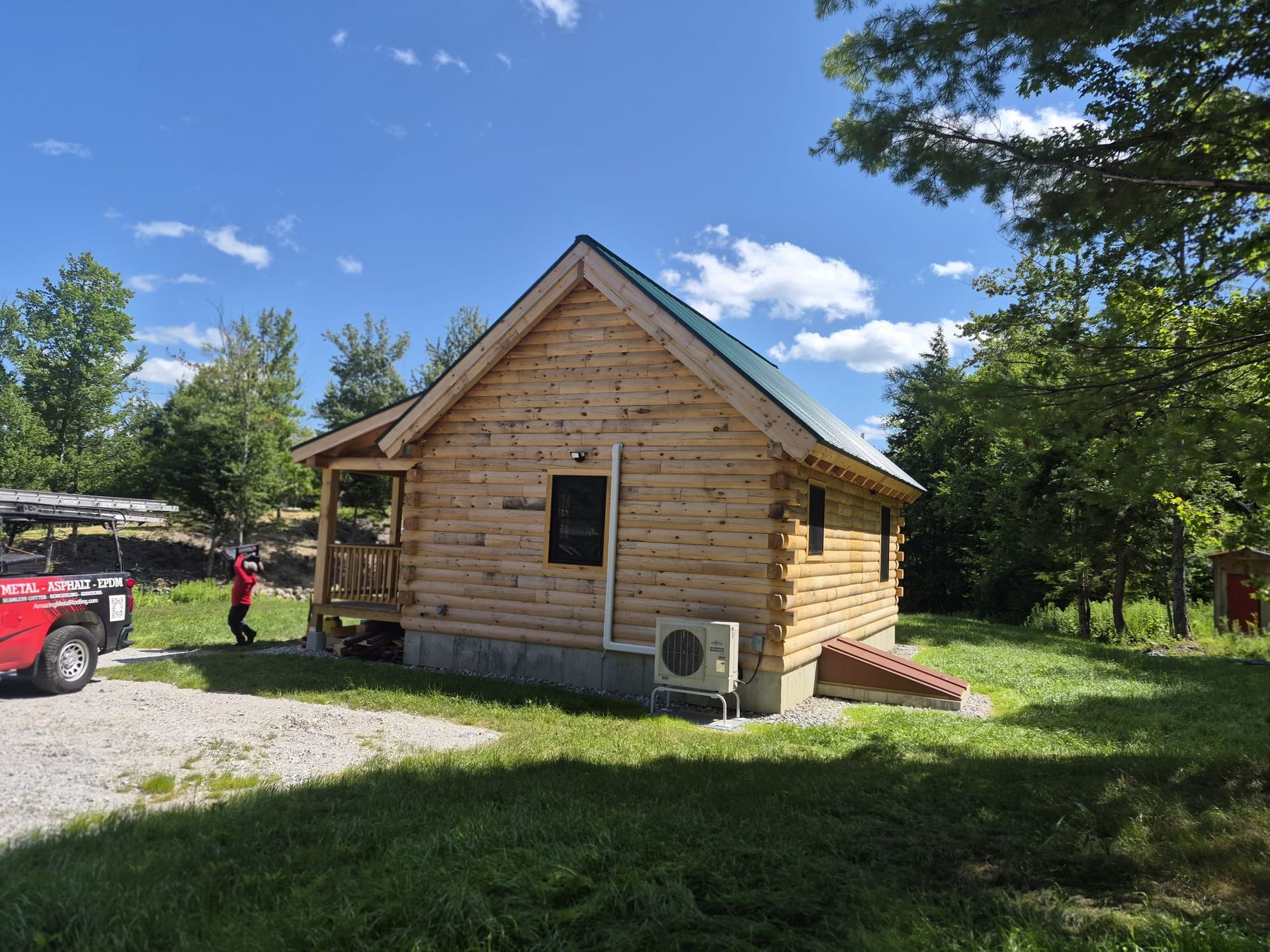 Wooden cabin with a gravel driveway, red truck, and trees under a bright blue sky
