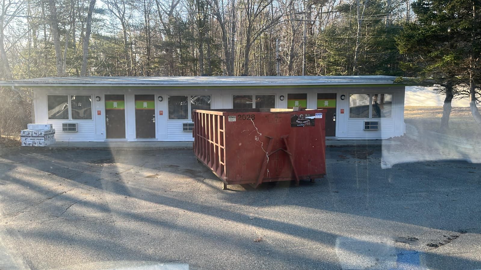 White roadside building with several doors and windows, a red dumpster in front, and trees behind.