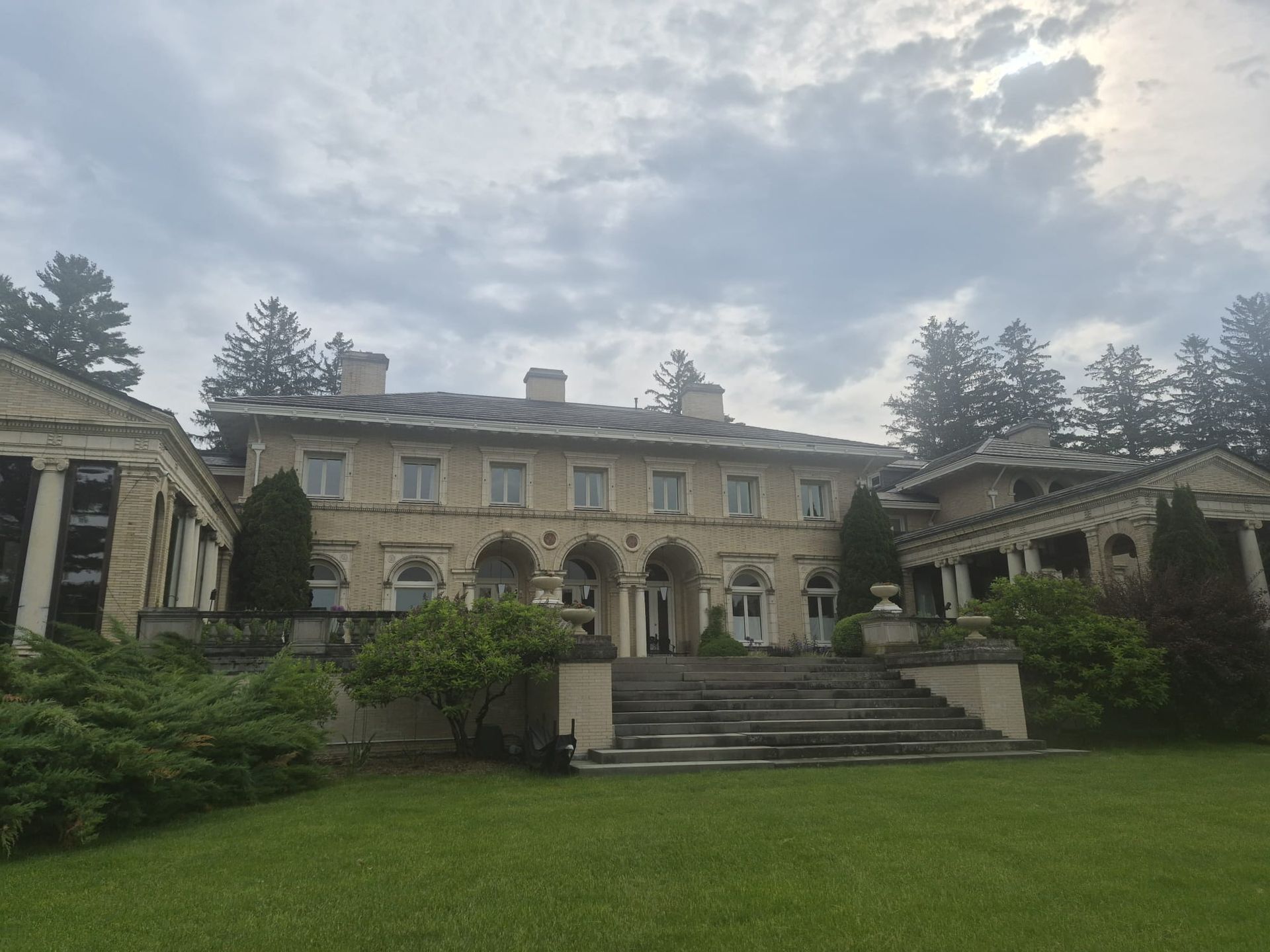 Large stone mansion with arched windows and a central staircase, viewed from a manicured lawn under a cloudy sky