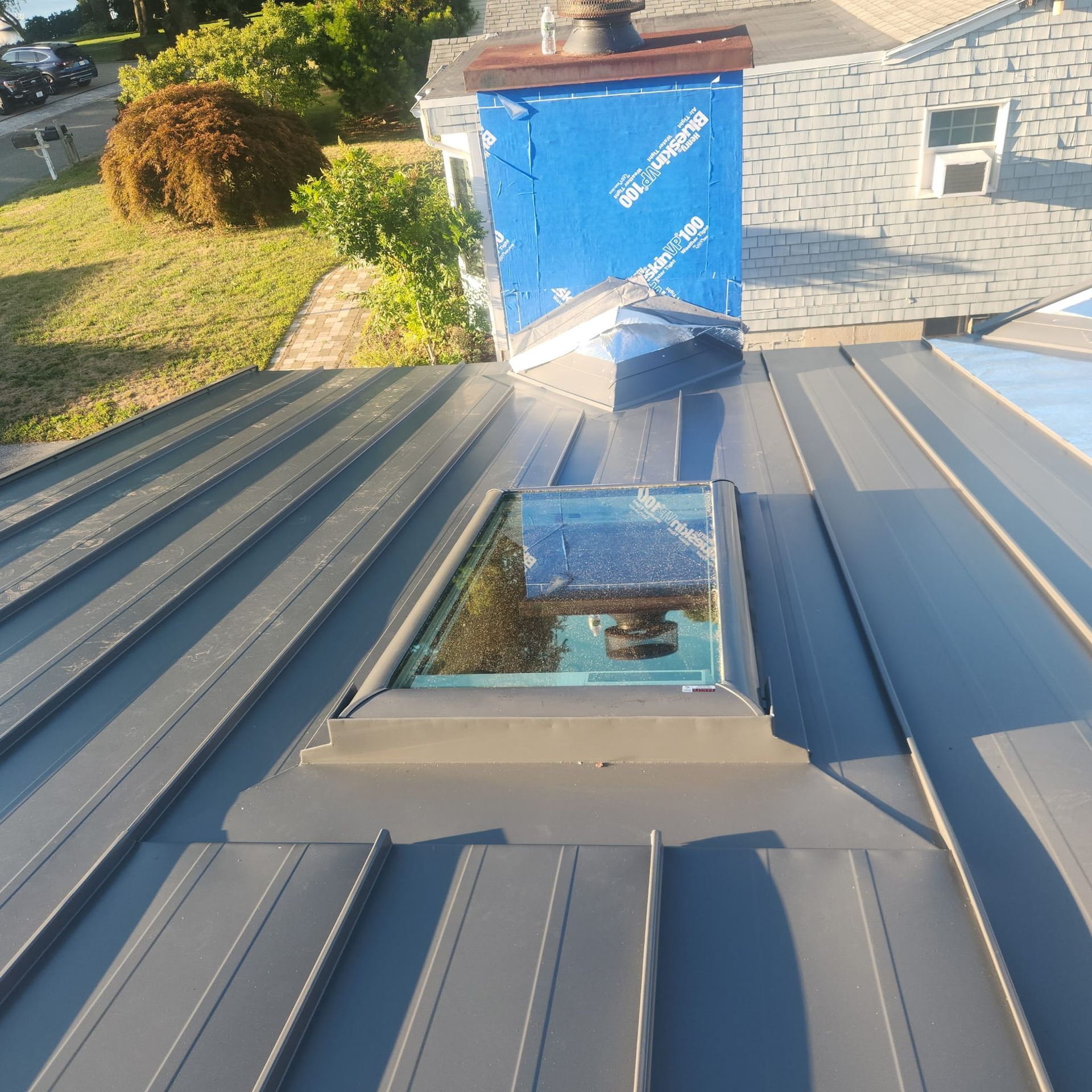 Gray metal roof with a rectangular skylight reflecting the sky, next to a blue tarp-covered structure.