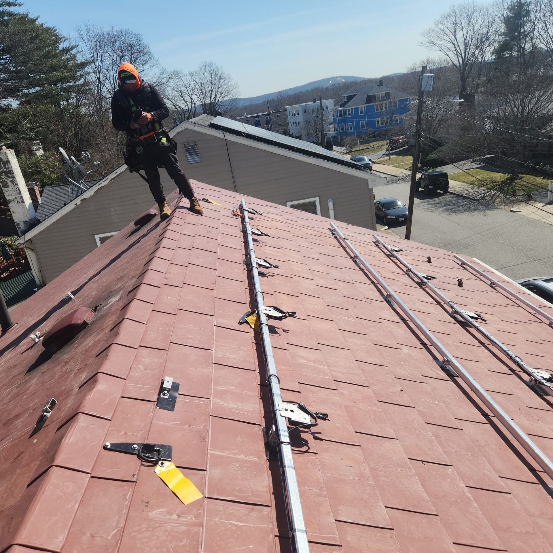 Worker on a red roof using safety gear, with houses and trees in the background.