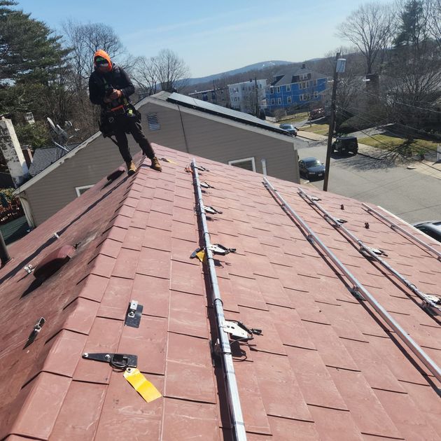 Worker on a red shingled roof inspecting repairs on a sunny residential street.