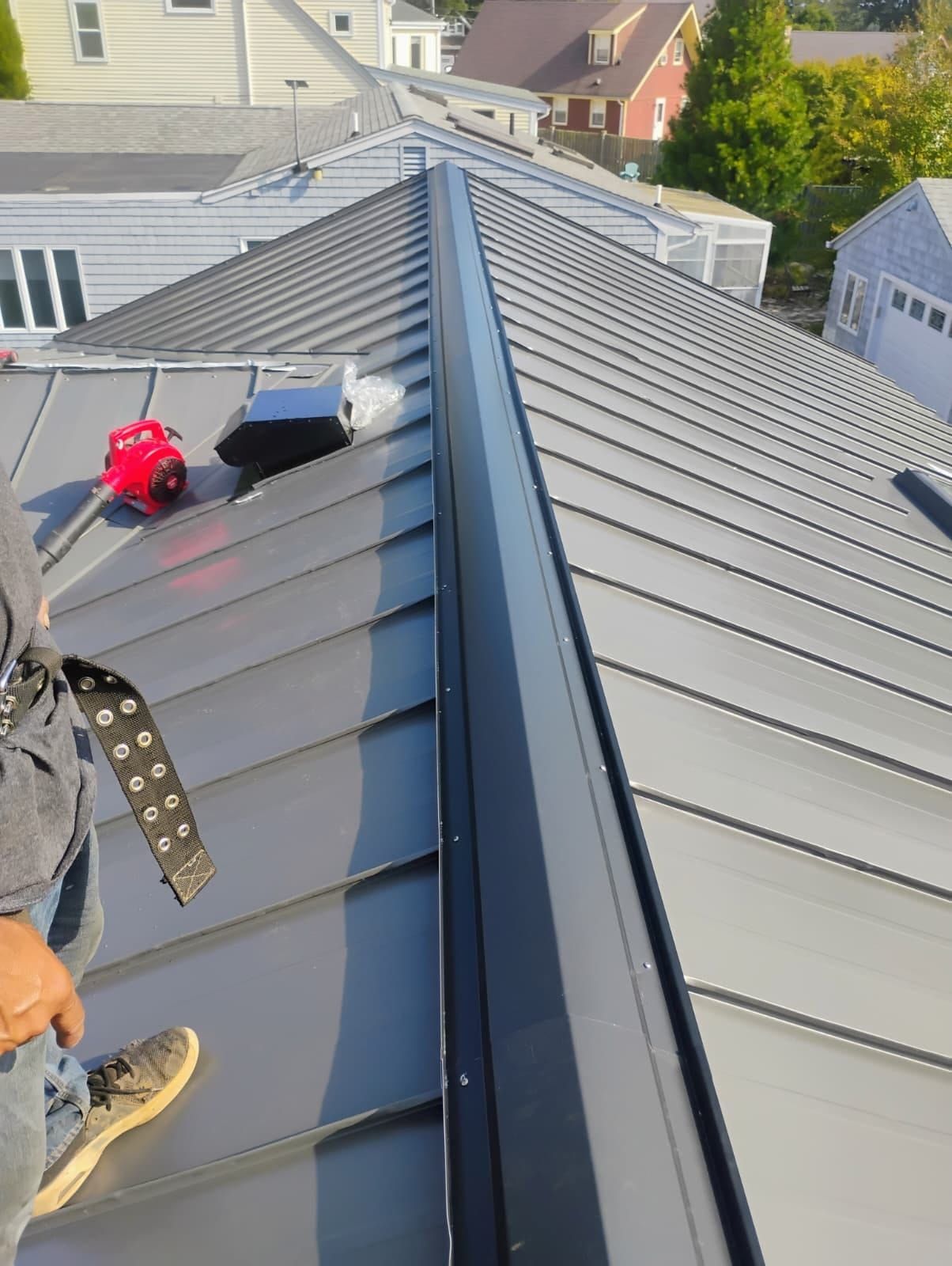 Roofers working on a gray metal roof ridge with tools and safety gear