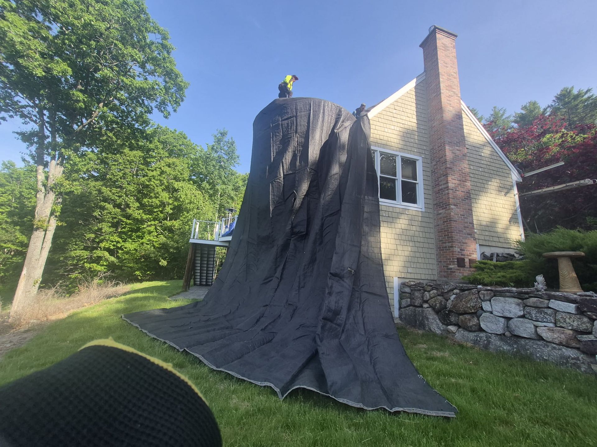 Black fabric draped over a tall tree stump in a sunny yard beside a stone-and-brick house.